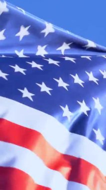 Açık mavi gökyüzüne karşı rüzgarda dalgalanan Amerikan bayrağının canlı yakın çekimi. Bu resim, vatanseverliği simgeleyen ABD bayrağının ikonik kırmızı, beyaz ve mavi renklerini yakalar.