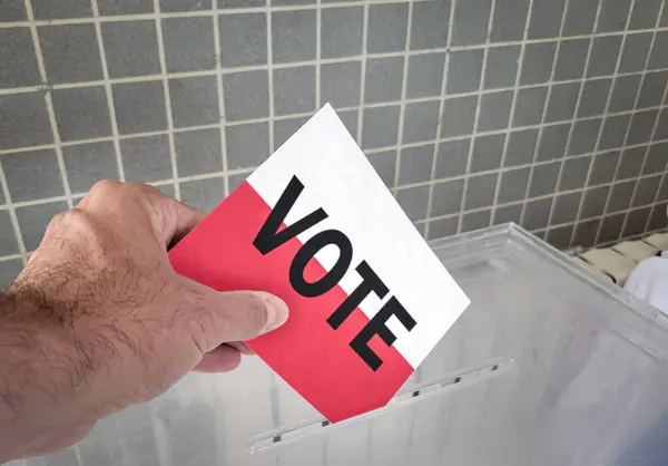 stock image A close-up photograph captures a hand placing a voting ballot with the word VOTE printed over the flag of Poland into a transparent ballot box. This image symbolizes the democratic process and the act