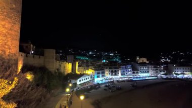 Costa Brava 'da popüler bir turizm merkezi olan Tossa de Mar' ın gece manzarası aydınlatılmış plajını ve tarihi şehir duvarlarını vurguluyor.