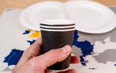 Elinde tek kullanımlık siyah bir kağıt bardak ve arka planda Avrupa Birliği 'nin haritası var. Tek kullanımlık plastik SUP Direktifi sembolize ediliyor