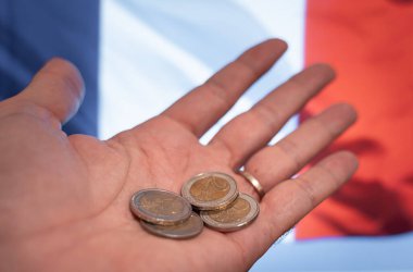 Arka planda Fransa bayrağı olan, yoksulluğu, mali krizi ve ekonomik zorlukları temsil eden birkaç avro senti gösteren bir el. Fransız hükümeti düşme riski altında