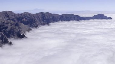 La Palma, Caldera de Taburiente, dağ zirvelerinden kalın bir bulut katmanıyla zaman çizelgesi, kaya oluşumları ve aşağıdaki yumuşak, beyaz bulutlar arasında çarpıcı bir kontrastlık yaratır..