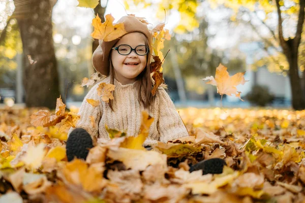 Happy cute brown-haired woman with Down\'s syndrome in a knitted sweater and stylish glasses throws bright foliage into the sky and laughs, the child enjoys time in a warm autumn park, happy childhood