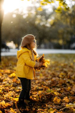 Down sendromlu, örgü kazaklı ve şık gözlüklü mutlu kahverengi saçlı kadın gökyüzüne parlak yapraklar fırlatıyor ve gülüyor, çocuk sıcak bir sonbahar parkında eğleniyor, mutlu bir çocukluk geçiriyor.