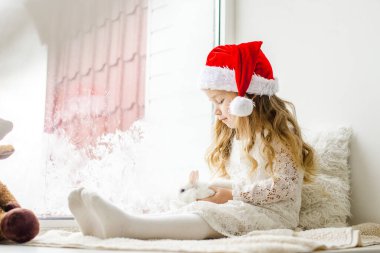 Penceresinde beyaz sevimli bir tavşanı olan küçük bir kız oturuyor. Çocuk pencereden dışarı bakar ve Noel Baba 'yı bekler. 2023 Tavşan sembolü