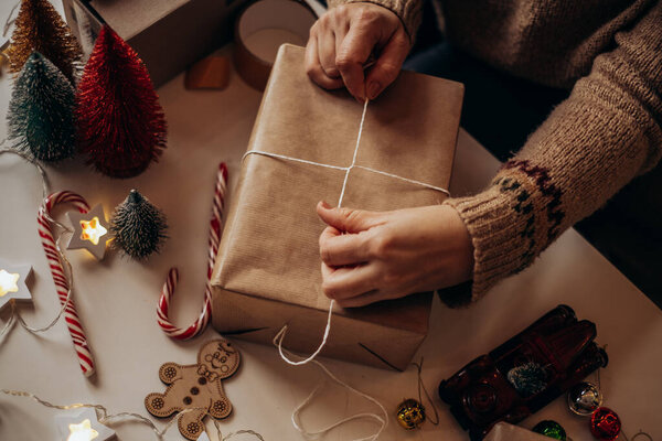 Женские руки заворачивают рождественские подарочные коробки, закрываются. Неподготовленные подарки на белом столе с элементами декора и элементами.