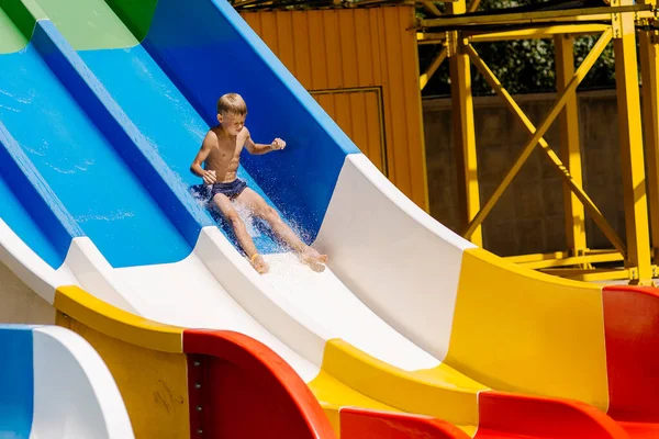 小さな男の子がウォータースライドを滑り降りて楽しんでいます 少年は夏にウォータースライダーを下った後プールで泳ぐ — ストック写真
