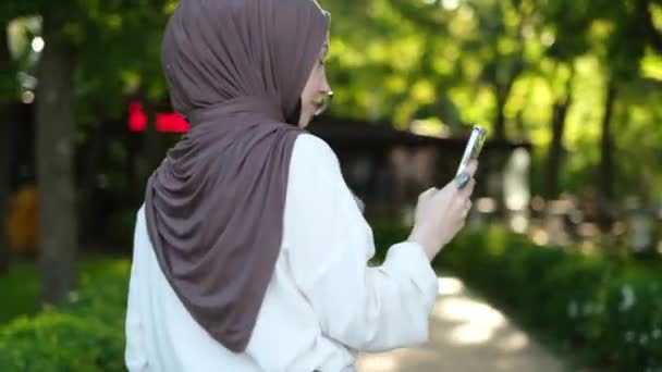 2人の若いイスラム教徒の女性がヒジャーブを着用し 公園を歩き スマートフォンを使用し 友人に会い 現代技術を満たし オンラインで写真を投稿 友人の再会の幸福 — ストック動画