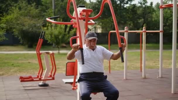 穿着运动裤和T恤衫的老年人抽动肌肉 在户外的铁模拟器上做健身锻炼 在一个特殊的场地上做运动 过着健康的生活 — 图库视频影像