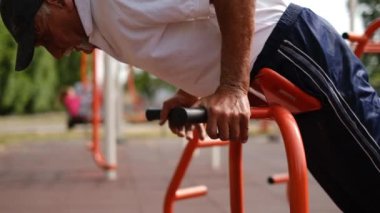 Eşofmanlı ve tişörtlü kıdemli bir adam kas pompalıyor, dışarıda demir simülatörde spor yapıyor, özel bir sitede spor yapıyor, sağlıklı bir hayat sürüyor.