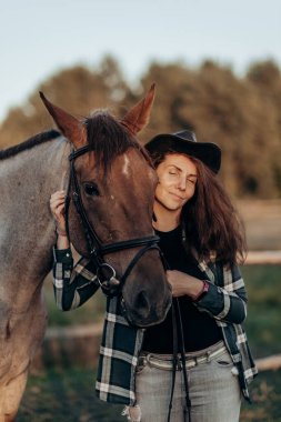 Gün batımında kahverengi atıyla oynayan şapkalı ve gömlekli güzel bir genç kadın. At biniciliği.