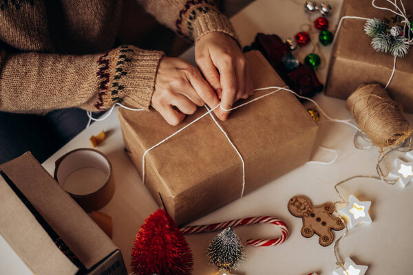 Женские руки заворачивают рождественские подарочные коробки, закрываются. Неподготовленные подарки на белом столе с элементами декора и элементами.