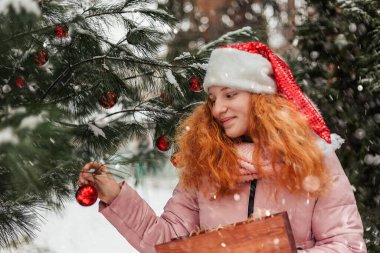 Noel Baba şapkalı kızıl saçlı bir kız karlı bir Noel ağacını Noel oyuncaklarıyla süslüyor.