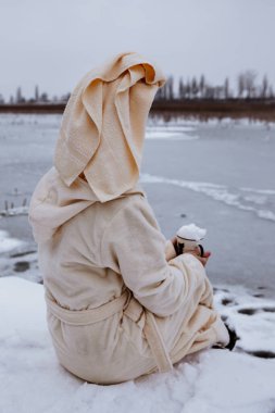 Kışın elinde bir kupa, dinlenme, kışın açık hava eğlencesi bornozla fotoğraf çekimi yaratıcı fotoğraf çekimi, kışın gölün kıyısında dinlenen bir kadın.