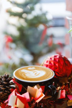 Çam kozalakları, kırmızı ve altın rengi tatil dekorları ve arka planda bulanık Noel ağacı ışıklarıyla çevrili ılık bir latte sanatı. Tatil promosyonları, sezonluk pazarlama ve indirimler için mükemmel..