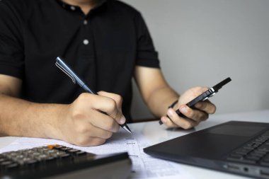 Vergi formunu kalem ve telefon kullanarak dolduran adam, beyaz masada hesap makinesi ve dizüstü bilgisayarla