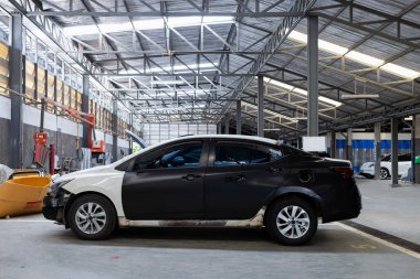 Sedan undergoing bodywork repair in auto workshop, with unfinished paint and replacement parts visible inside garage.