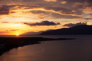 Bir sahil kasabasının üzerinde nefes kesici bir gün batımı. Güneş suyun üzerine sıcak bir ışık saçıyor. Manzara, arka planda dağlık bir arazi ve sakin bir koy içeriyor.,
