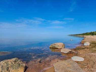 Küçük çakıl taşları ve daha büyük kayalarla kayalar durgun, berrak mavi sulara, uzak, yemyeşil bir ormana ve berrak mavi gökyüzüne, tüylü, beyaz bulutlara götürür..