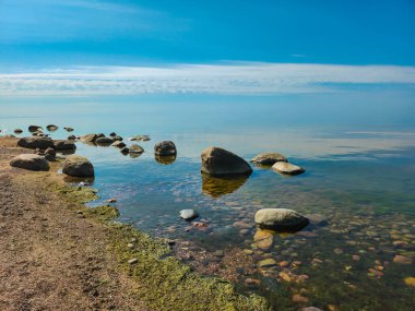 Görüntü büyük taşları ve mavi suyu olan kayalık bir sahili gösteriyor. Su berrak ve gökyüzünü yansıtıyor. Taşlar farklı boyutlarda ve şekillerde ve sahile saçılmışlar..