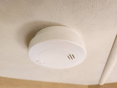 Beyaz bir plastik duman dedektörü, dokulu bir tavana monte edilmiş, merkezinde küçük bir delik olan yuvarlak ve beyaz bir arka planda üç küçük yarık var..