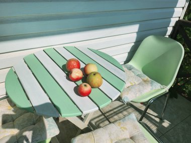 Görüntü, yeşil, dört elmalı beyaz çizgili ve yanında minderleri olan iki sandalyeli yuvarlak ahşap bir masa gösteriyor. Masa ve sandalyeler beyaz arka planda izole edilmiştir..