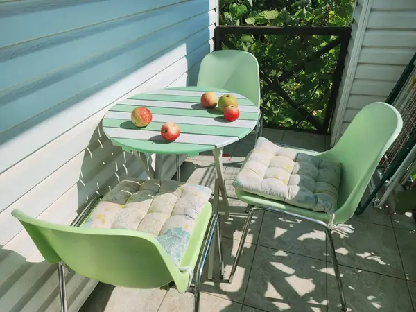 Bahçede rahat bir oturma alanı. Yastıklı üç yeşil sandalyeli yuvarlak bir masa. Masada birkaç elma var. Arkaplan yeşil yapraklarla bulanıklaşmış..
