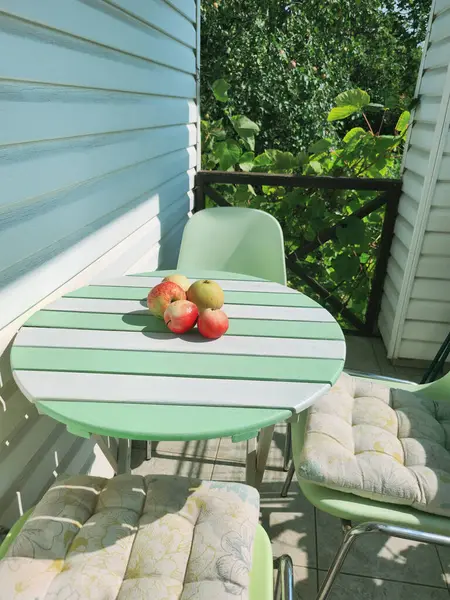 Yeşil ve beyaz çizgili yuvarlak bir masa, dört sandalye ve sakin bir bahçe terasında olgun kırmızı elmalar. yemyeşil duvar, saksı bitkileri, arka planda açık mavi gökyüzü..