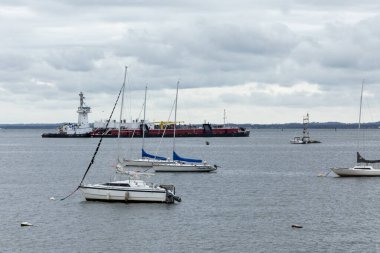 Perth Amboy, New Jersey - October 1, 2024: A Tugboat pushes a barge through the Arthur Kill in Perth Amboy. Sailboats from the Raritan Yacht Club are seen in the foreground. clipart