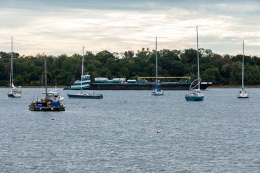 Perth Amboy, New Jersey - October 1, 2024: A Tugboat pushes a barge through the Arthur Kill in Perth Amboy. Sailboats from the Raritan Yacht Club are seen in the foreground. clipart