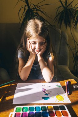 Kız resimlerle resim çiziyor. Yaratıcı gelişim ve mutlu çocukluk kavramı. İnternette bir öğretmenden resim dersleri alıyorum. Sanat terapisi. Masanın üzerinde bir palet boya ve kağıt var.