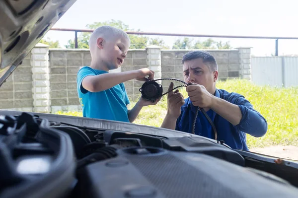 A mechanic dad teaches his son how to change the timing belt and car cooling pump at home. Child holds a pump in his hand. Maintenance of the car according to the regulations. New parts