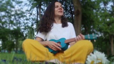 Parkta çimlerin üzerinde oturan kız ukulele çalıyor. Dört telli enstrüman çalmayı öğreniyorum. Halka açık bir parkta rahatlama. Müzik terapisi, psikoloji, meditasyon, rahatlama, rahatlama.