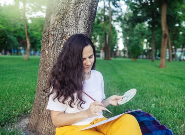 Sanat terapisi. Kadın parkta çizim yapıyor. Kız sırtı ağaca dönük oturur ve mesafeye bakar. Çizim yardımı ile sinir sisteminin yenilenmesi. Rahatlama ve meditasyon durumu kavramı