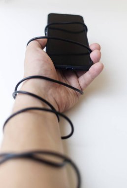 Telefon bağımlılığının üstesinden gelmek. Erkek eli akıllı telefon şarj aletinin etrafına dolanmış. Akıllı telefon bağımlılığının kavramsal fotoğrafı. Normal bir hayat yaşama yeteneği kaybı. Dijitalleşmenin olumsuz etkisi