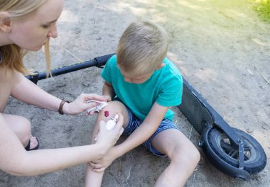 Çocuk elektrikli scooter sürerken düşmüş. Koruyucu giysi olmadan e-scooter kullanmak. Pille çalışan araçlar kullanıldığında yaralanma riski artıyor. Annem diz sakatlığı için ilk yardım yapıyor.
