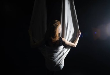 Sinek yogası. Siyah arka planda beyaz hamakta oturan kız hava akrobasisi gösteriyor. Jimnastik, sirk, kubbe altında. Işık ve gölge oyunu tiyatral bir performans hissi yaratır. Spor aktivitesi