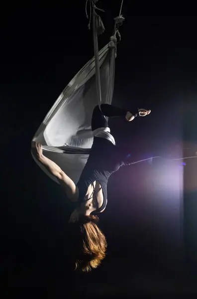Fly yoga. Girl in a white hammock on a black background shows aerial acrobatics. Gymnastics, circus, under dome. Play of light and shadow creates feeling of a theatrical performance. Sports activity