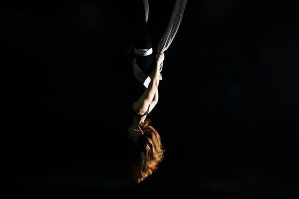 Fly yoga. Girl in a white hammock on a black background shows aerial acrobatics. Gymnastics, circus, under dome. Play of light and shadow creates feeling of a theatrical performance. Sports activity