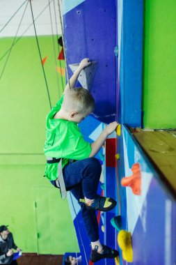 Çocuklar için kaya tırmanışı. Yeşil gömlekli sarışın çocuk duvara tırmanıyor. Kayalık sınıfı. Güvenli aktiviteler ve yarışmalar için yapay yol. Dağcılık, güvenlik sistemi, özel ayakkabılar.