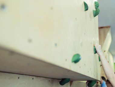 Çocuklar için kaya tırmanışı. Yeşil gömlekli sarışın çocuk duvara tırmanıyor. Kayalık sınıfı. Güvenli aktiviteler ve yarışmalar için yapay yol. Dağcılık, güvenlik sistemi, özel ayakkabılar.