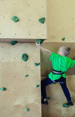 Çocuklar için kaya tırmanışı. Yeşil gömlekli sarışın çocuk duvara tırmanıyor. Kayalık sınıfı. Güvenli aktiviteler ve yarışmalar için yapay yol. Dağcılık, güvenlik sistemi, özel ayakkabılar.