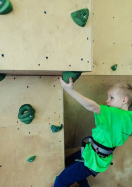 Çocuklar için kaya tırmanışı. Yeşil gömlekli sarışın çocuk duvara tırmanıyor. Kayalık sınıfı. Güvenli aktiviteler ve yarışmalar için yapay yol. Dağcılık, güvenlik sistemi, özel ayakkabılar.