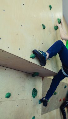 Çocuklar için kaya tırmanışı. Yeşil gömlekli sarışın çocuk duvara tırmanıyor. Kayalık sınıfı. Güvenli aktiviteler ve yarışmalar için yapay yol. Dağcılık, güvenlik sistemi, özel ayakkabılar.