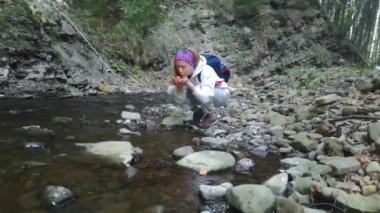 Turistler nehirden su içer. Dağ nehrinden su içen kız yolcuların yakın çekimi. Taşlar, orman manzarası, küçük gölet. Kaynatılmamış su içmekten kaynaklanan bakteriyel enfeksiyon ve ishal tehlikesi