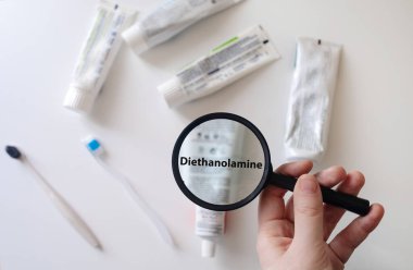 Dangerous toothpaste ingredient diethanolamine. Checking the composition of toothpaste with a magnifying glass against the background of many tubes clipart