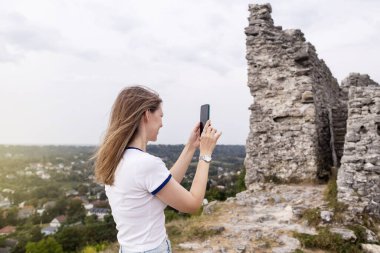 Genç bir kadın telefonuyla dağın tepesinden şehrin güzel bir manzarasının fotoğrafını çekiyor. Akıllı telefondan fotoğraflar ve videolar. Seyahat blogcusu ve etkileyicisi