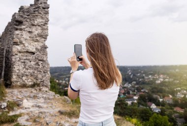 Genç bir kadın telefonuyla dağın tepesinden şehrin güzel bir manzarasının fotoğrafını çekiyor. Akıllı telefondan fotoğraflar ve videolar. Seyahat blogcusu ve etkileyicisi