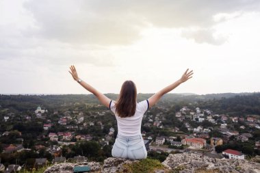Genç bir kadın, elleri bir kazanan gibi havada bir dağda oturuyor. Görülen manzaradan bir özgürlük ve memnuniyet jesti. Yukarıdan güzel bir şehir manzarası. Tatil, dağlarda yürüyüş, aktif yaşam tarzı.