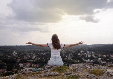 Genç bir kadın, elleri bir kazanan gibi havada bir dağda oturuyor. Görülen manzaradan bir özgürlük ve memnuniyet jesti. Yukarıdan güzel bir şehir manzarası. Tatil, dağlarda yürüyüş, aktif yaşam tarzı.